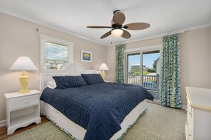 A bedroom with a bed, two bedside tables with lamps, a ceiling fan, a window with blinds, and a view of the outside. - At Ease: Ocean views, short walk to the beach, close to fishing pier. (Nags Head)