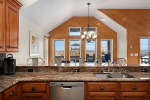 A kitchen with granite countertops, wooden cabinets, a built-in dishwasher, and a view of the beach through large windows.