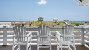 En balkong med tre vita Adirondack-stolar som vetter mot en strand med träkonstruktioner och en klar himmel.
