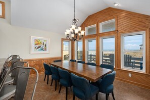 A dining area with a large wooden table, blue upholstered chairs, and a chandelier. - The Sandbox: Oceanfront, private pool, hot tub, rec & theater rooms, elevator! (Kill Devil Hills)