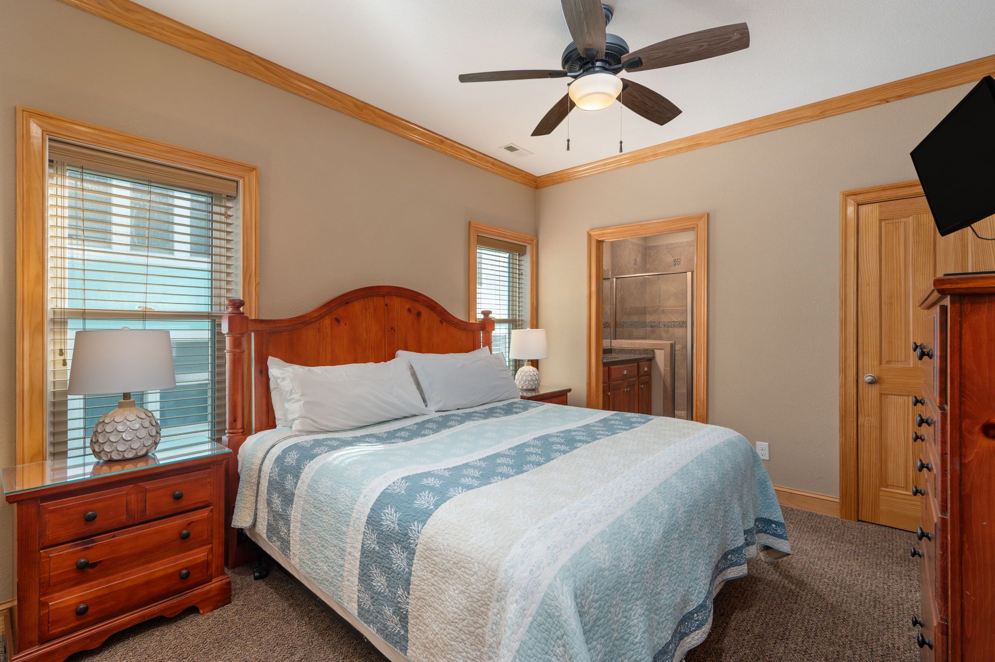 Une chambre à coucher avec un lit, des tables de chevet en bois, un ventilateur de plafond, un miroir et une porte.