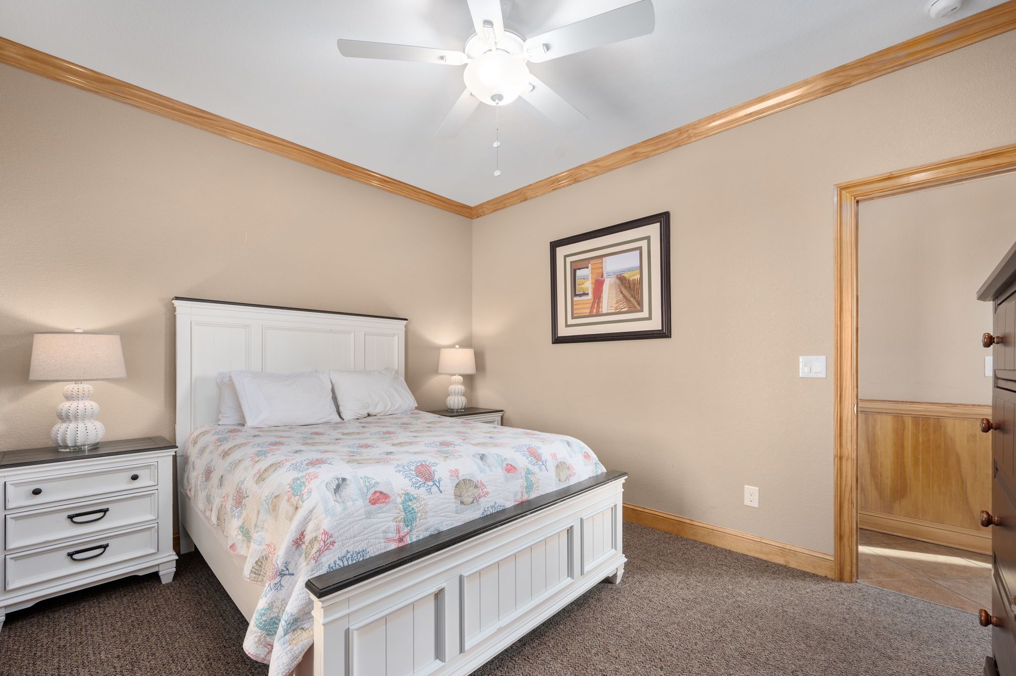 Quarto com cama, mesas de cabeceira, ventilador de teto, quadro na parede e porta de madeira.