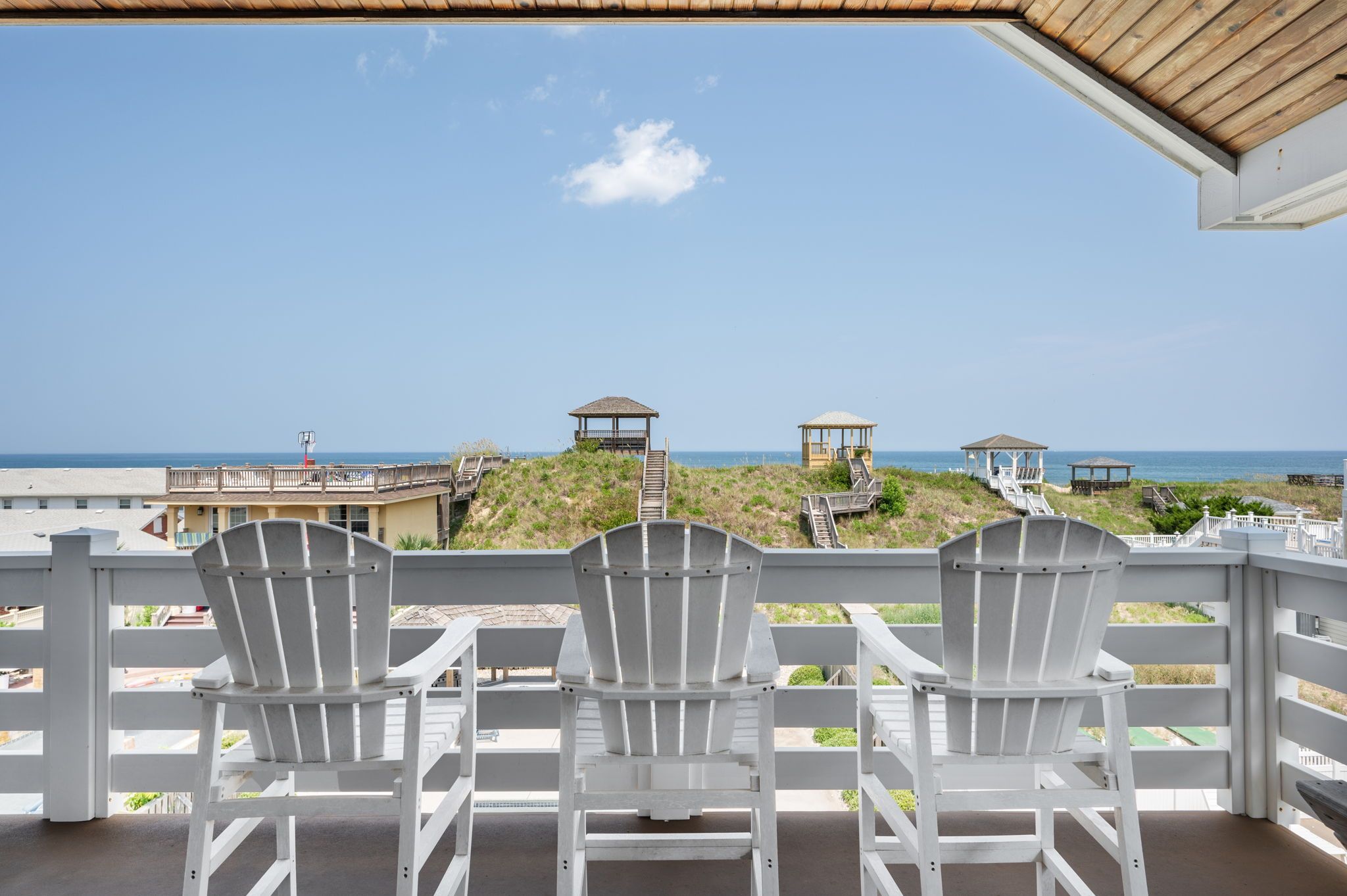A balcony with three white Adirondack chairs facing a beach with wooden structures and a clear sky.