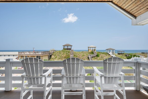 A balcony with three white Adirondack chairs facing a beach with wooden structures and a clear sky.