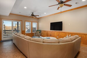 A living room with a large L-shaped sectional sofa, a flat-screen TV mounted on the wall, and a ceiling fan. - The Sandbox: Oceanfront, private pool, hot tub, rec & theater rooms, elevator! (Kill Devil Hills)
