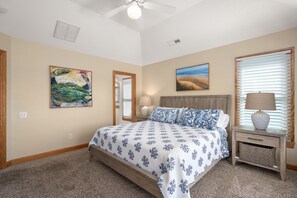 A bedroom with a bed, a nightstand, a lamp, a painting, and a window with blinds. - Sunset Shack: Soundfront, great sunsets, hot tub, community pool & tennis courts (Nags Head)