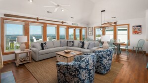 A spacious living area with a large sectional sofa, a coffee table, and patterned chairs. There are multiple windows offering a view of the ocean. - Sunset Shack: Soundfront, great sunsets, hot tub, community pool & tennis courts (Nags Head)
