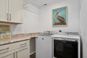 A laundry room with a Maytag washer and dryer, a painting of a pelican, and a sign that reads 'HAPPENS BEACH'.