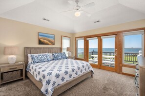 A bedroom with a large bed, a nightstand, a lamp, a ceiling fan, and a view of the ocean through a sliding glass door.