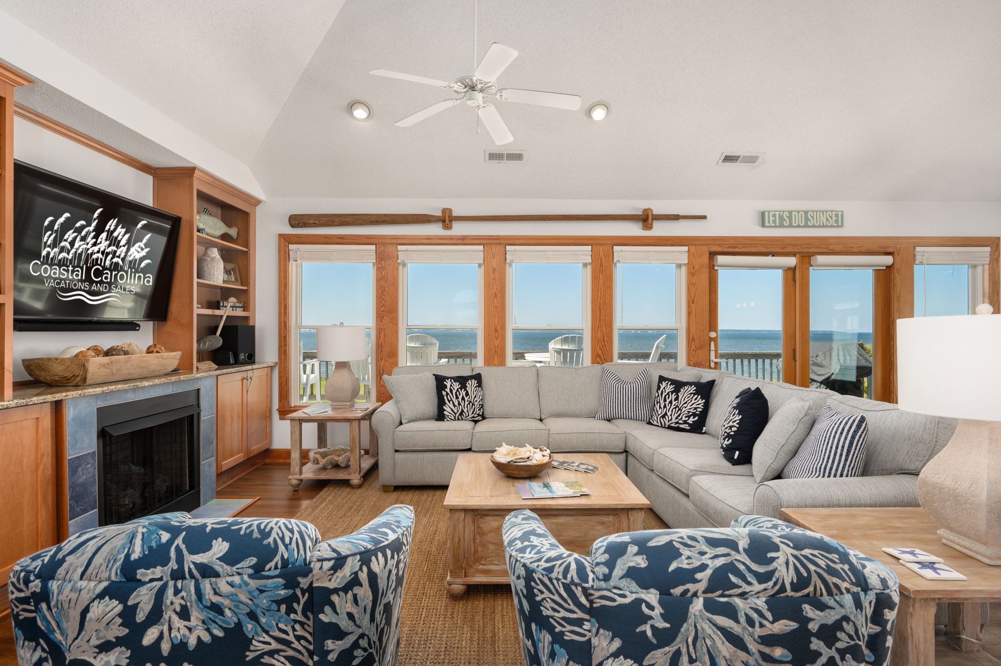 A spacious living room with a large window overlooking the ocean, a fireplace, and a television displaying 'Coastal Carolina Vacations and Sales'.