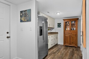 A kitchen with a stainless steel refrigerator, white cabinets, and a wooden cabinet with books. - Sunset Shack: Soundfront, great sunsets, hot tub, community pool & tennis courts (Nags Head)