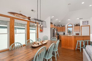 A modern kitchen with wooden cabinets, a central island, and a dining area with a wooden table and chairs. - Sunset Shack: Soundfront, great sunsets, hot tub, community pool & tennis courts (Nags Head)