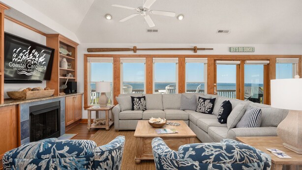 A spacious living room with a large window overlooking the ocean, a fireplace, and a television displaying 'Coastal Carolina Vacations and Sales'. - Sunset Shack: Soundfront, great sunsets, hot tub, community pool & tennis courts (Nags Head)