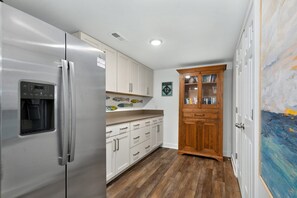 A kitchen with a stainless steel refrigerator, white cabinets, a wooden cabinet with books, and a painting on the wall. - Sunset Shack: Soundfront, great sunsets, hot tub, community pool & tennis courts (Nags Head)