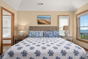 A bedroom with a large bed, two nightstands, a headboard, a painting, and a window with a view. - Sunset Shack: Soundfront, great sunsets, hot tub, community pool & tennis courts (Nags Head)