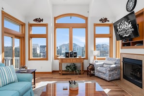 A spacious living room with large windows, a fireplace, and a television displaying 'Coastal Carolina Vacations and Sales'. - Piggy's Paradise-Oceanside, community pool, private hot tub, short walk to the beach! (Nags Head)