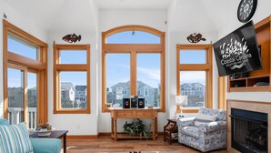 Un salón espacioso con ventanas amplias, una chimenea y un televisor mostrando 'Coastal Carolina Vacations and Sales'.