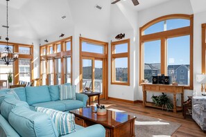 A spacious living room with a large sectional sofa, wooden flooring, and large windows offering a view of the neighborhood. - Piggy's Paradise-Oceanside, community pool, private hot tub, short walk to the beach! (Nags Head)