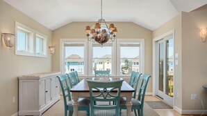A dining area with a chandelier, white cabinets, teal chairs, and large windows with a view of houses. - Beach Brum: Oceanside, private heated saltwater pool, hot tub, elevator, rec room with pool table (Nags Head)