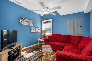 Uma sala de estar com um sofá vermelho, uma TV de tela plana, um ventilador de teto e obras de arte nas paredes.