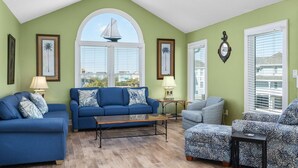 Una sala de estar con un sofá azul, un sillón gris y un sillón estampado. Hay dos lámparas, una ventana con persianas y una decoración de un velero en la pared.