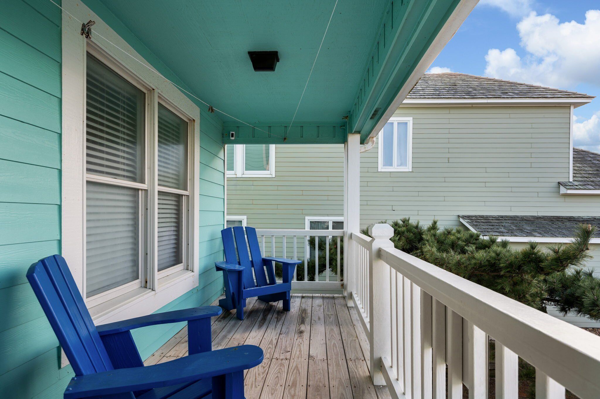 A porch with two blue Adirondack chairs, a white railing, and a view of a house with a green roof.