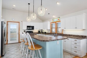 A kitchen with white cabinets, a granite countertop, and a bar area with stools. - Moonstruck: Semi-oceanfront, private pool, hot tub & beach is across the street! (Nags Head)