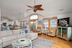 A spacious living room with a large sofa, wicker chairs, a ceiling fan, and a television displaying "Coastal Carolina Vacations and Sales".