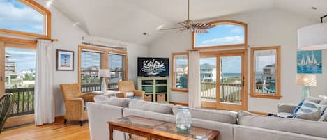 A spacious living room with a large window overlooking the sea, a ceiling fan, and a television on the wall.