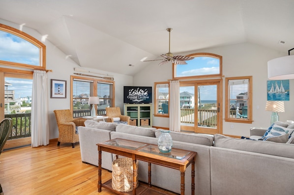 A spacious living room with a large window overlooking the sea, a ceiling fan, and a television on the wall.