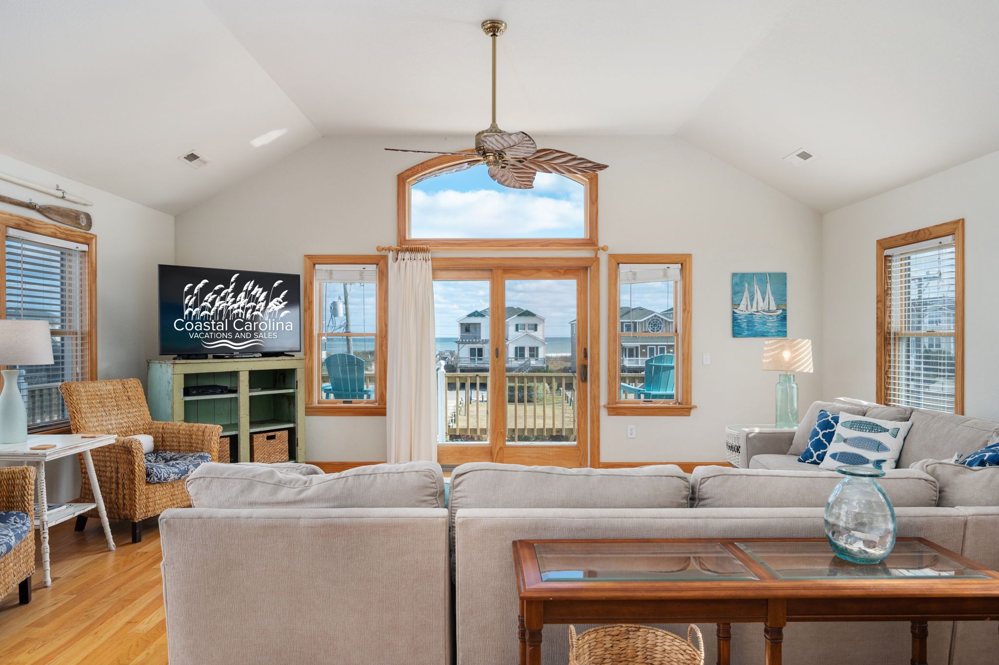 A spacious living room with a large window, a ceiling fan, and a television displaying 'Coastal Carolina Vacations and Sales'.