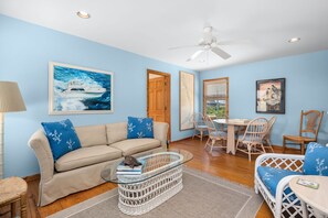 A living room with a beige sofa, a glass coffee table, wicker chairs, a dining table, and artwork on the walls.