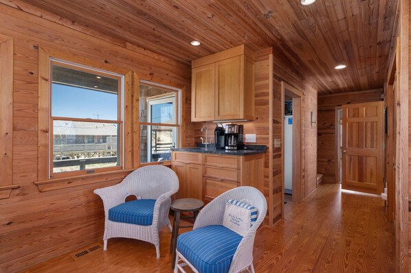 A wooden cabin interior with a kitchen, two wicker chairs, a small table, and a view through a window.
