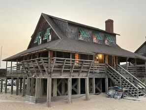 A wooden building with a balcony and stairs, situated on a sandy area.