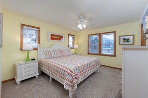 Une chambre à coucher comprenant un lit, deux tables de chevet, un ventilateur de plafond et une fenêtre avec des stores.