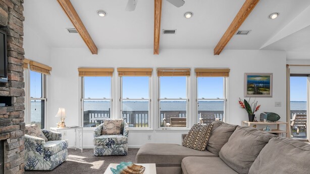 Een woonkamer met een stenen open haard, grote ramen met uitzicht op zee, een beige bank en een gestreept fauteuil.