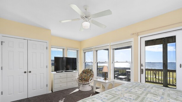 A bedroom with a bed, a television, a chair, and a view of the ocean through large windows.