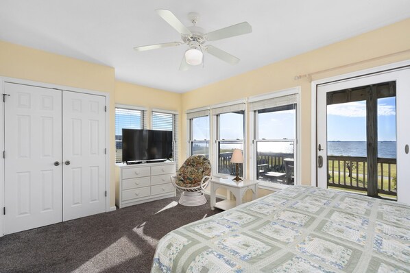 A bedroom with a bed, a television, a chair, and a view of the ocean through large windows.
