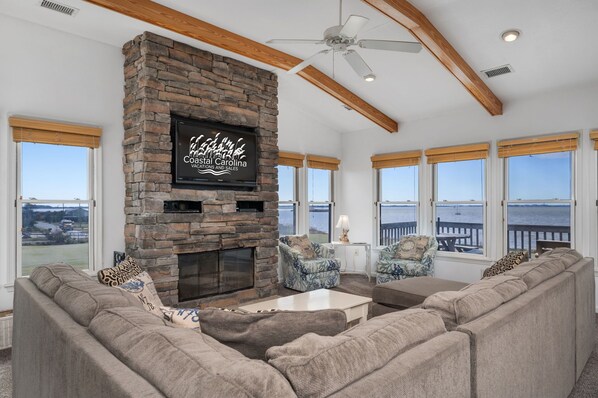 A spacious living room with a stone fireplace, large windows with ocean views, and a ceiling fan.