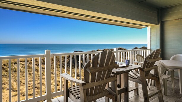 Eine Holzterrasse mit Stühlen und einem Tisch mit Blick auf einen Strand.