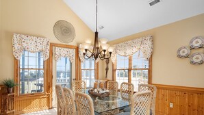 A dining area with a glass table surrounded by wicker chairs, a chandelier, and decorative plates on the wall. - Wedges 13: Soundside, 13th fairway view, great community pool and tennis courts. ⛳☀️ (Nags Head)