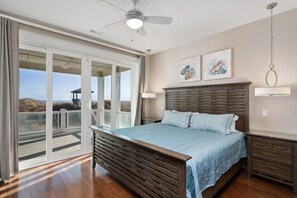 A bedroom with a large bed, a ceiling fan, a nightstand, and a view through a sliding glass door to an outdoor deck. - Sunset Beach House: Great views, private pool, hot tub, pool table & elevator (Nags Head)