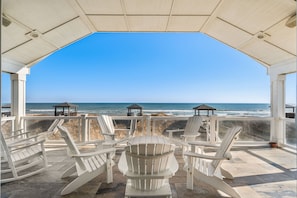A covered outdoor seating area with white Adirondack chairs and a central fire pit, overlooking the ocean.