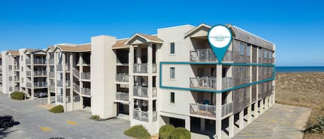 A multi-story hotel with balconies, a parking lot, and a view of the ocean.