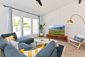 A modern living room with a large flat-screen TV, grey sofas, a coffee table, and a view of a cityscape through large windows.