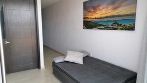 Um quarto com um sofá cinzento, um quadro de uma paisagem ao pôr do sol e uma porta fechada.