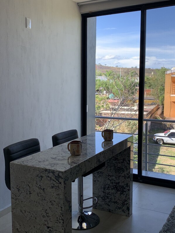 A dining area with a marble table, two chairs, and two decorative cups. A large window offers a view of a residential area with houses and a car. - Comfortable Modern Stay by Hidden Gem Vacation Homes (Zapotlanejo)
