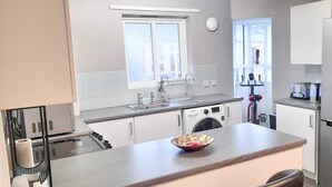 Une cuisine moderne équipée d’un four encastré, d’un lave-linge, d’un réfrigérateur et d’une fenêtre laissant entrer la lumière naturelle.