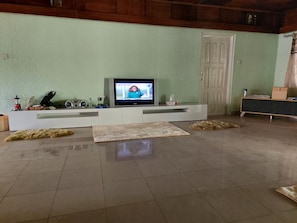 Una sala de estar con una televisión sobre un mueble blanco, un aparador de madera y suelo de baldosas.