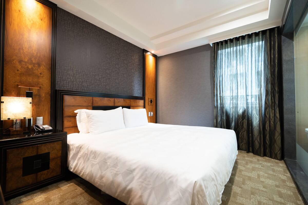 Habitación de hotel con una cama grande, una mesita de noche con lámpara, cabecera de madera y una ventana con cortinas.